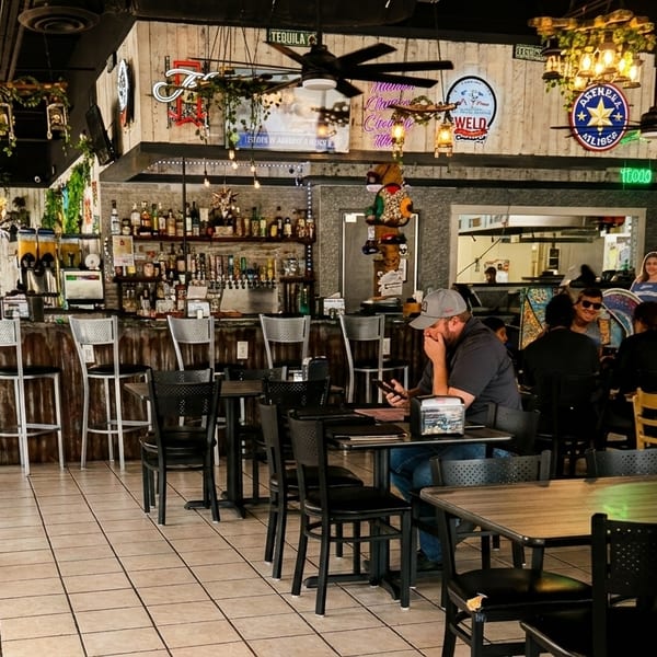 A vibrant restaurant interior with a bar, tables, and patrons, featuring colorful decor and hanging plants.