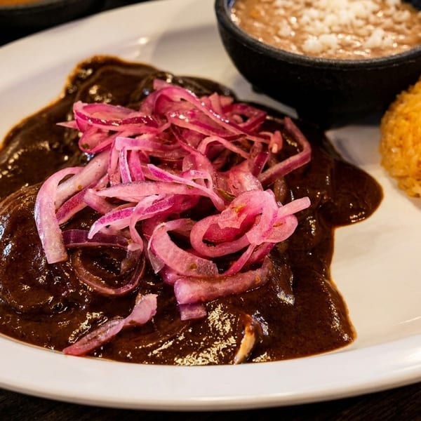 Chicken mole with pickled red onions, a side of rice, and refried beans on a white plate.