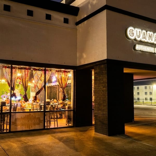 Exterior of Guanatos Grub at night, with a lit sign and patrons visible through the windows.
