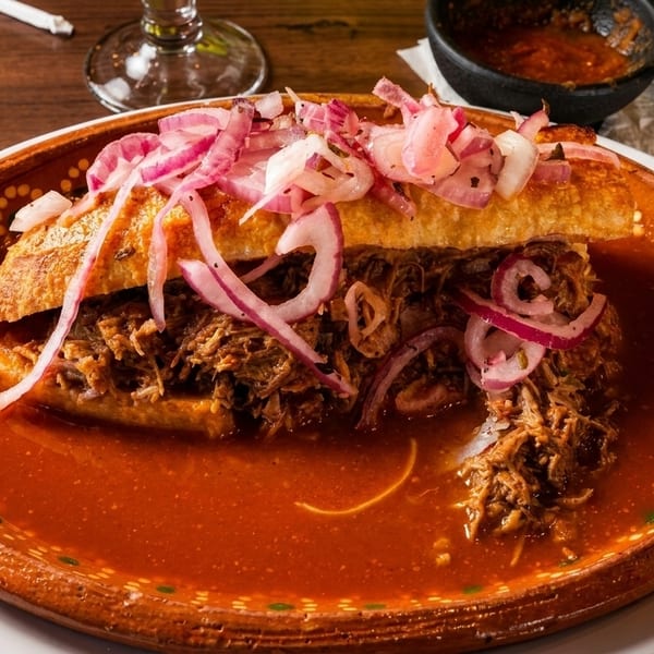 Mexican torta ahogada with shredded meat and pickled red onions in a red sauce.