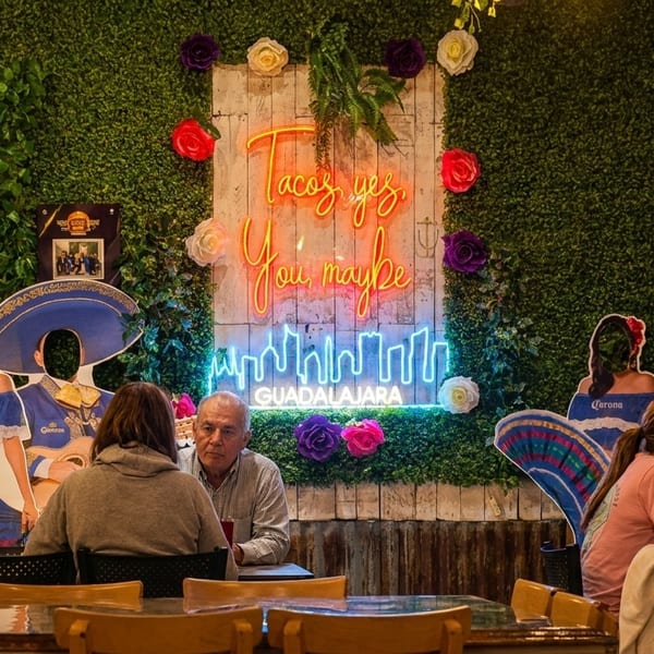Diners seated at tables in a restaurant with a neon "Tacos yes, You maybe" sign on a green wall.