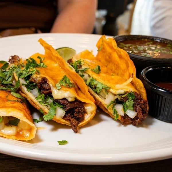 Three birria tacos with cheese, cilantro, and onions, served with dipping sauces on a white plate.