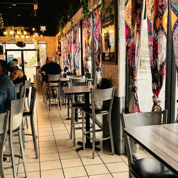 Restaurant interior with patrons at a bar and high-top tables, colorful curtains, and string lights.
