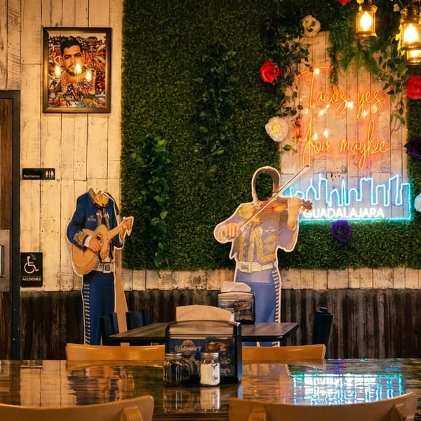 Restaurant interior with mariachi cutouts, a neon sign, and wooden tables under warm hanging lights.