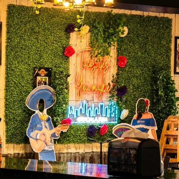 A vibrant restaurant interior with a neon "Taco yes, You maybe" sign on a green wall.